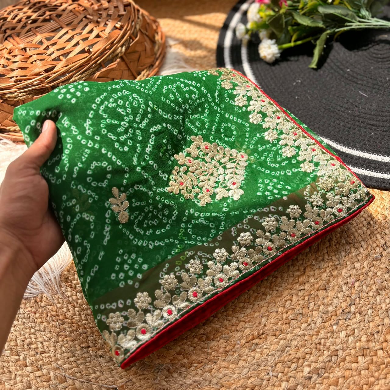 Deep Green Bandhani Georgette Saree With Gota Patti Work 1 A person holds a green Bandhani Georgette saree with gold Gota Patti embroidery on a rustic background.