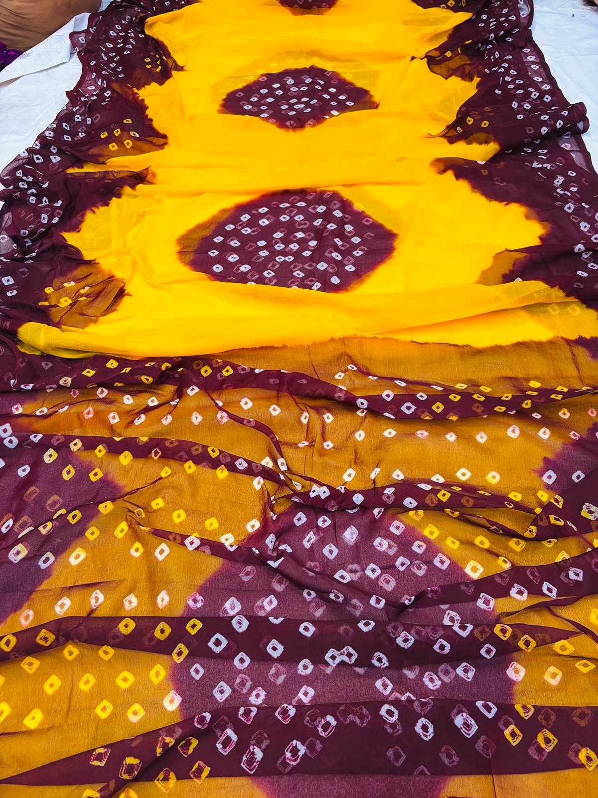 Nazneen Chiffon Hand Tie Dye Bandhani Saree 1 A close-up of a Nazneen Chiffon saree featuring a striking hand tie dye Bandhani pattern in yellow and maroon.