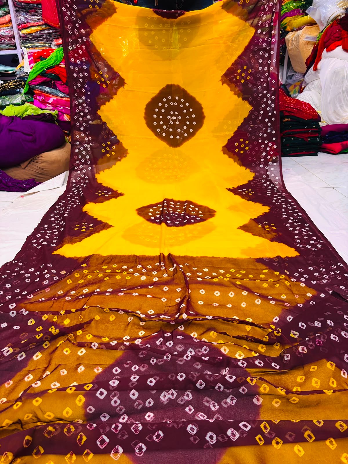 Nazneen Chiffon Hand Tie Dye Bandhani Saree 4 A close-up of a Nazneen Chiffon saree featuring a striking hand tie dye Bandhani pattern in yellow and maroon.