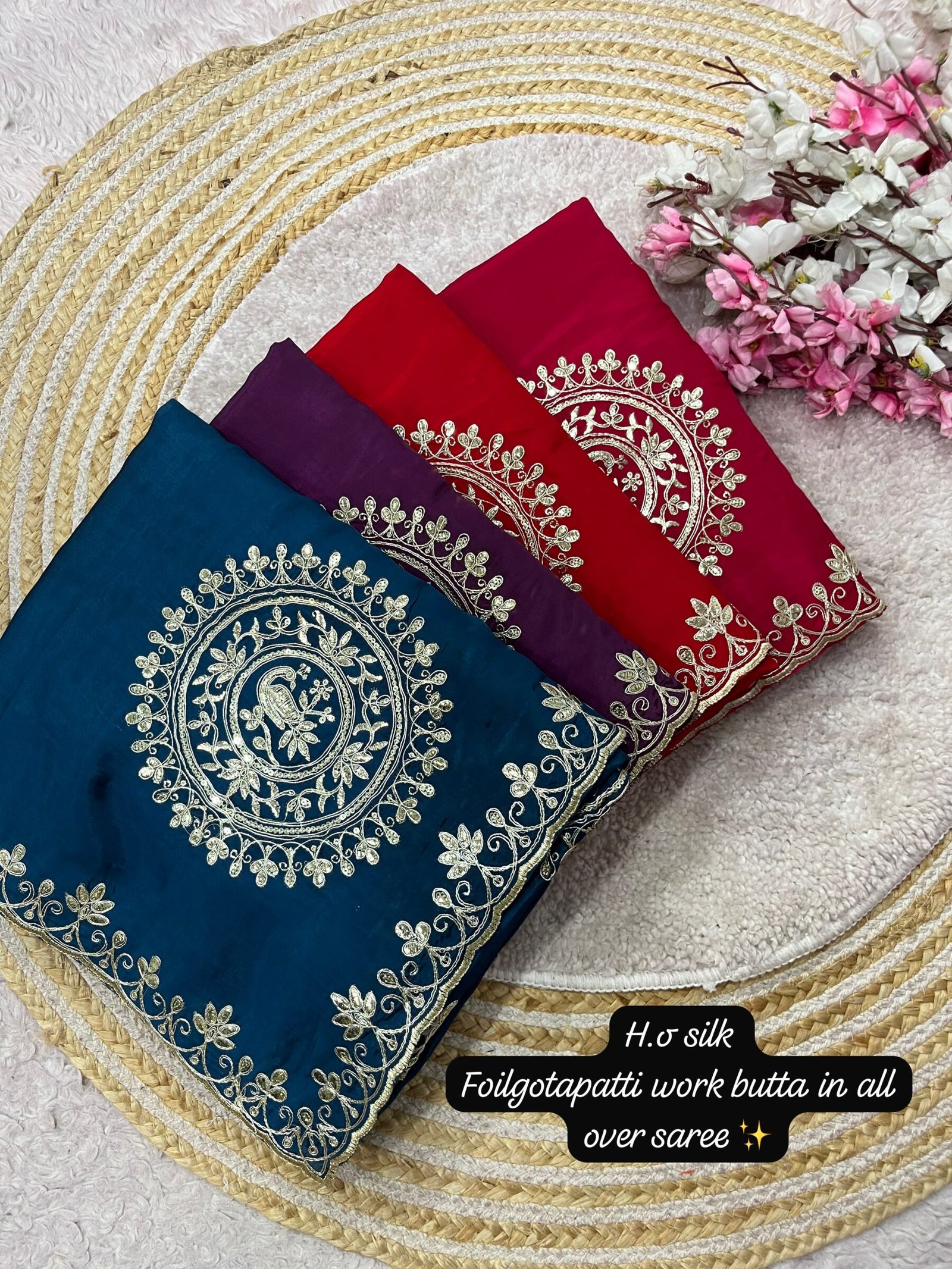 H.O Silk Gota Patti Saree 1 Four folded H.O silk sarees in blue, purple, and red displayed on a round woven mat with white and pink flowers.