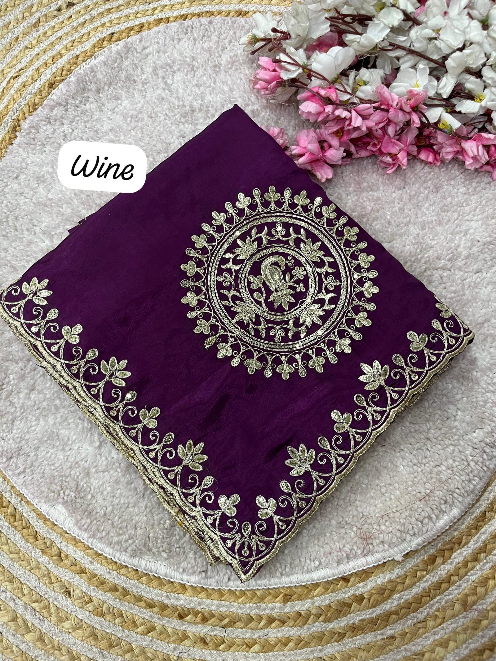 H.O Silk Gota Patti Saree 2 Four folded H.O silk sarees in blue, purple, and red displayed on a round woven mat with white and pink flowers.