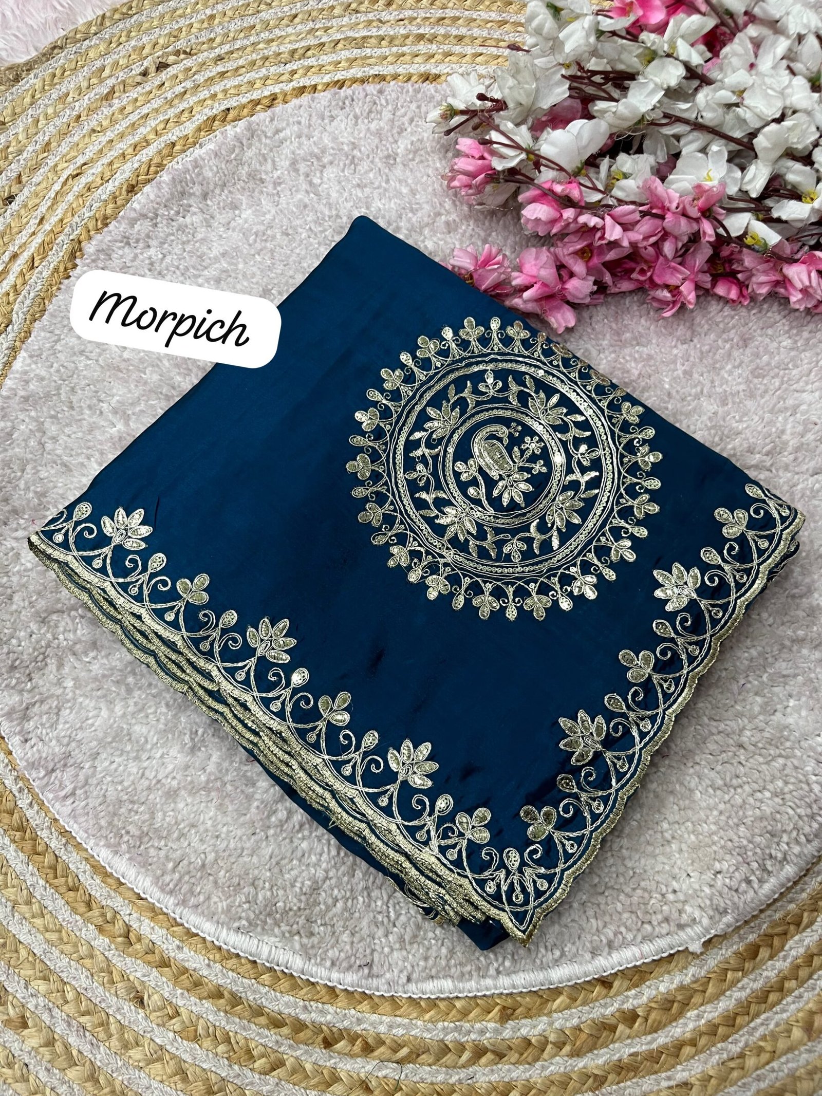 H.O Silk Gota Patti Saree 3 Four folded H.O silk sarees in blue, purple, and red displayed on a round woven mat with white and pink flowers.