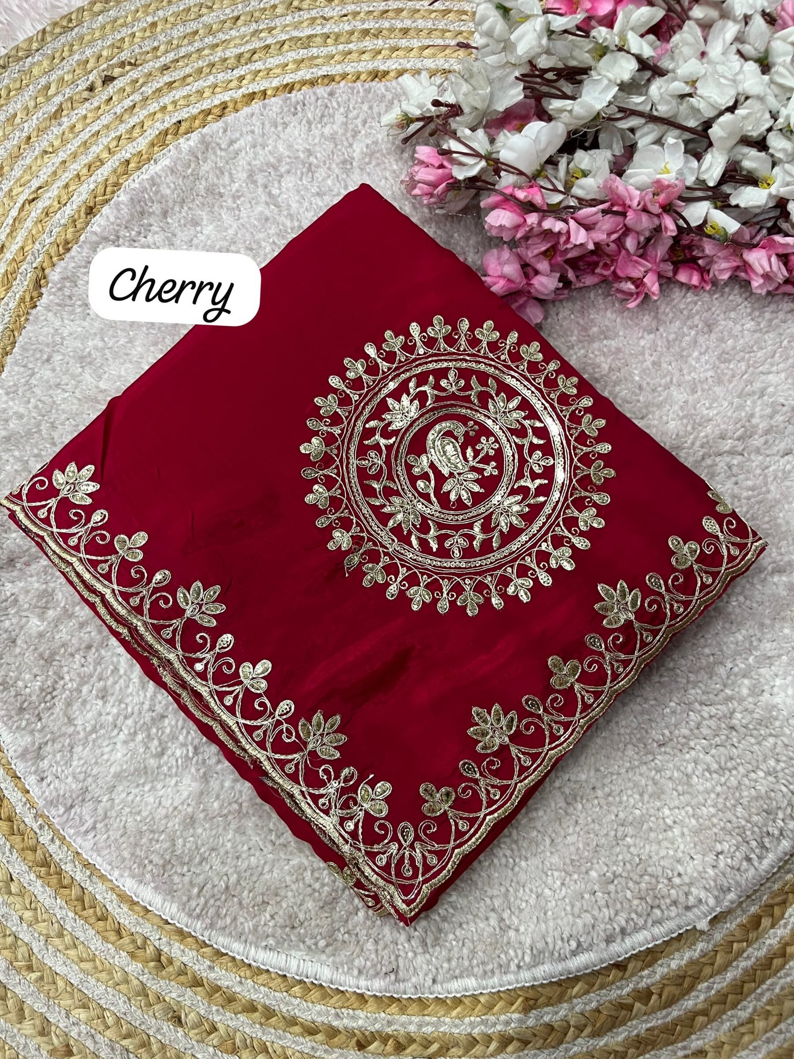 H.O Silk Gota Patti Saree 4 Four folded H.O silk sarees in blue, purple, and red displayed on a round woven mat with white and pink flowers.