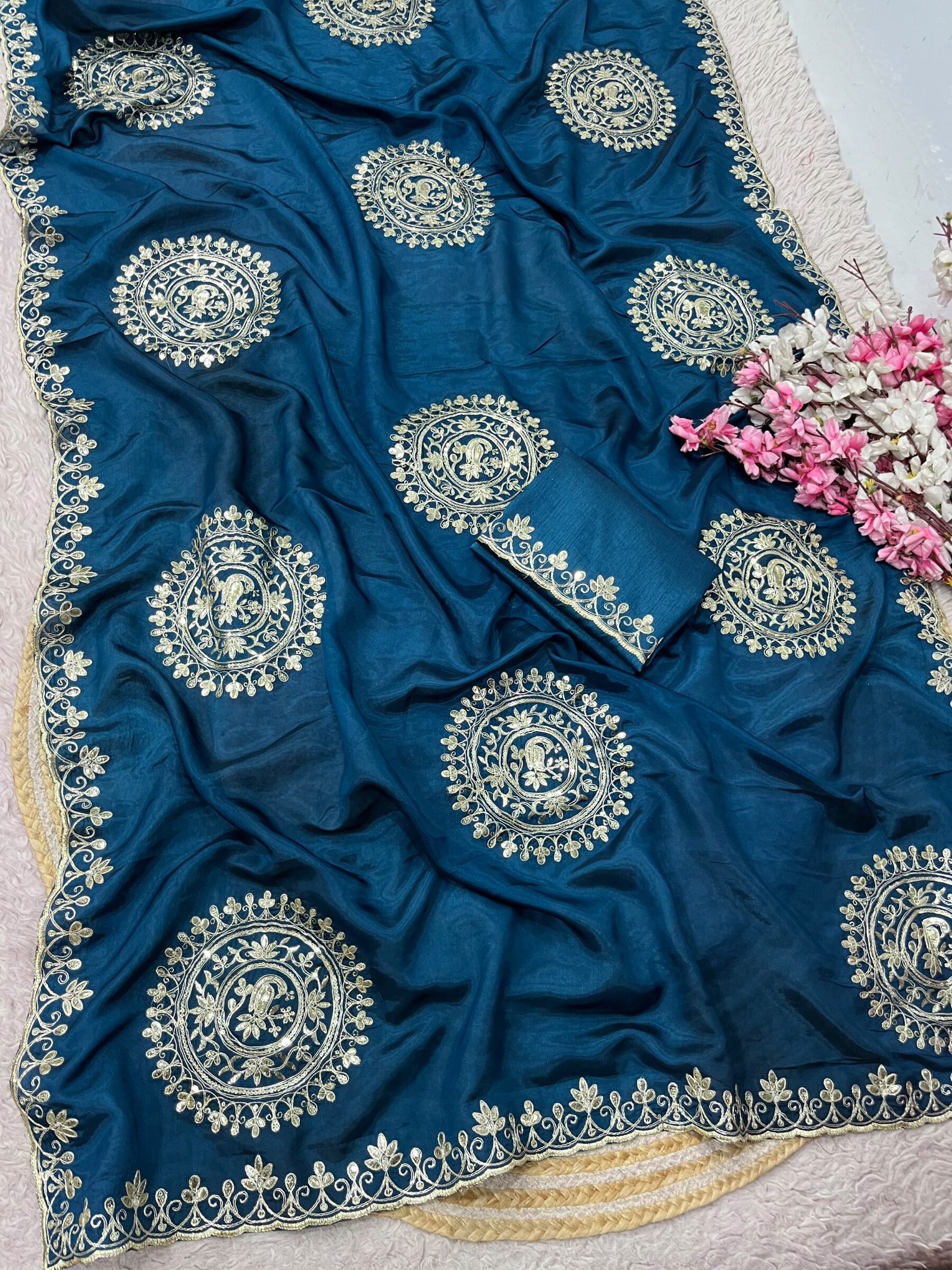 H.O Silk Gota Patti Saree 9 Four folded H.O silk sarees in blue, purple, and red displayed on a round woven mat with white and pink flowers.