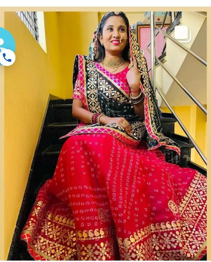 A woman wearing a vibrant red and black Kota Doriya Bandhej lehenga with heavy Gota Patti embroidery while sitting on a staircase.