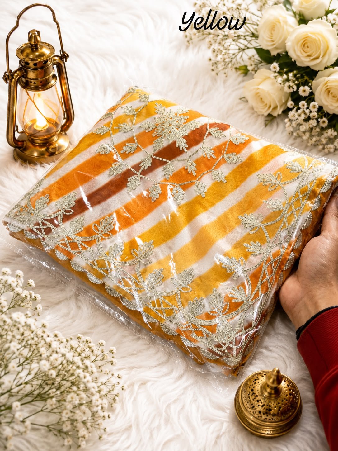 Chinon Leheriya Saree with Kacchi Gotapatti Work 3 Three folded Leheriya sarees in vibrant colors with Kacchi Gotapatti work placed near a vintage lantern and white roses.