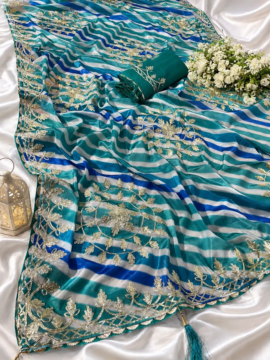 Chinon Leheriya Saree with Kacchi Gotapatti Work 5 Three folded Leheriya sarees in vibrant colors with Kacchi Gotapatti work placed near a vintage lantern and white roses.