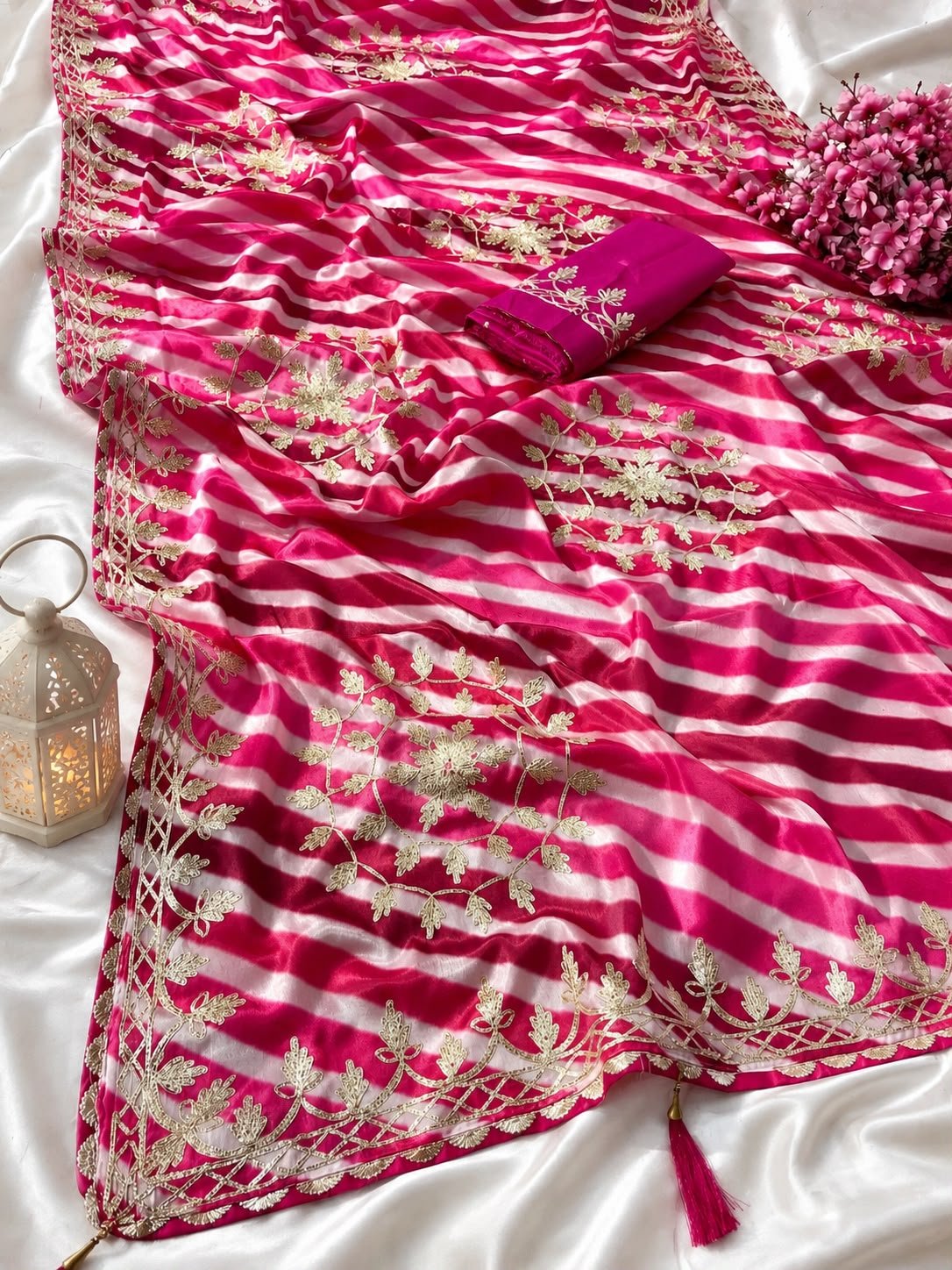 Chinon Leheriya Saree with Kacchi Gotapatti Work 6 Three folded Leheriya sarees in vibrant colors with Kacchi Gotapatti work placed near a vintage lantern and white roses.