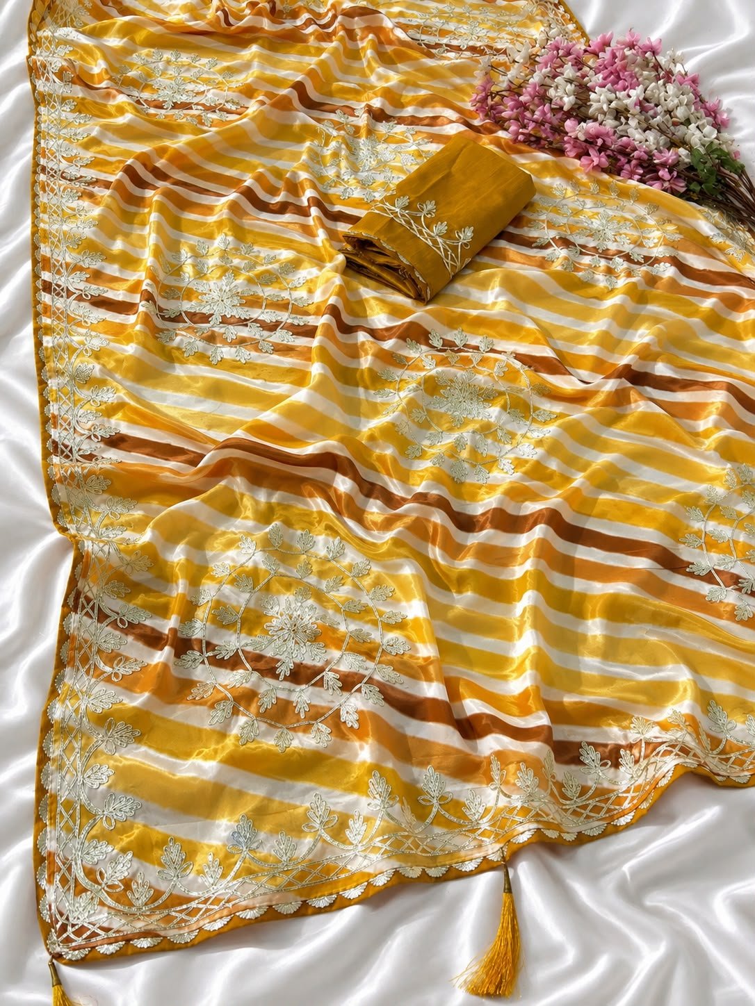 Chinon Leheriya Saree with Kacchi Gotapatti Work 7 Three folded Leheriya sarees in vibrant colors with Kacchi Gotapatti work placed near a vintage lantern and white roses.