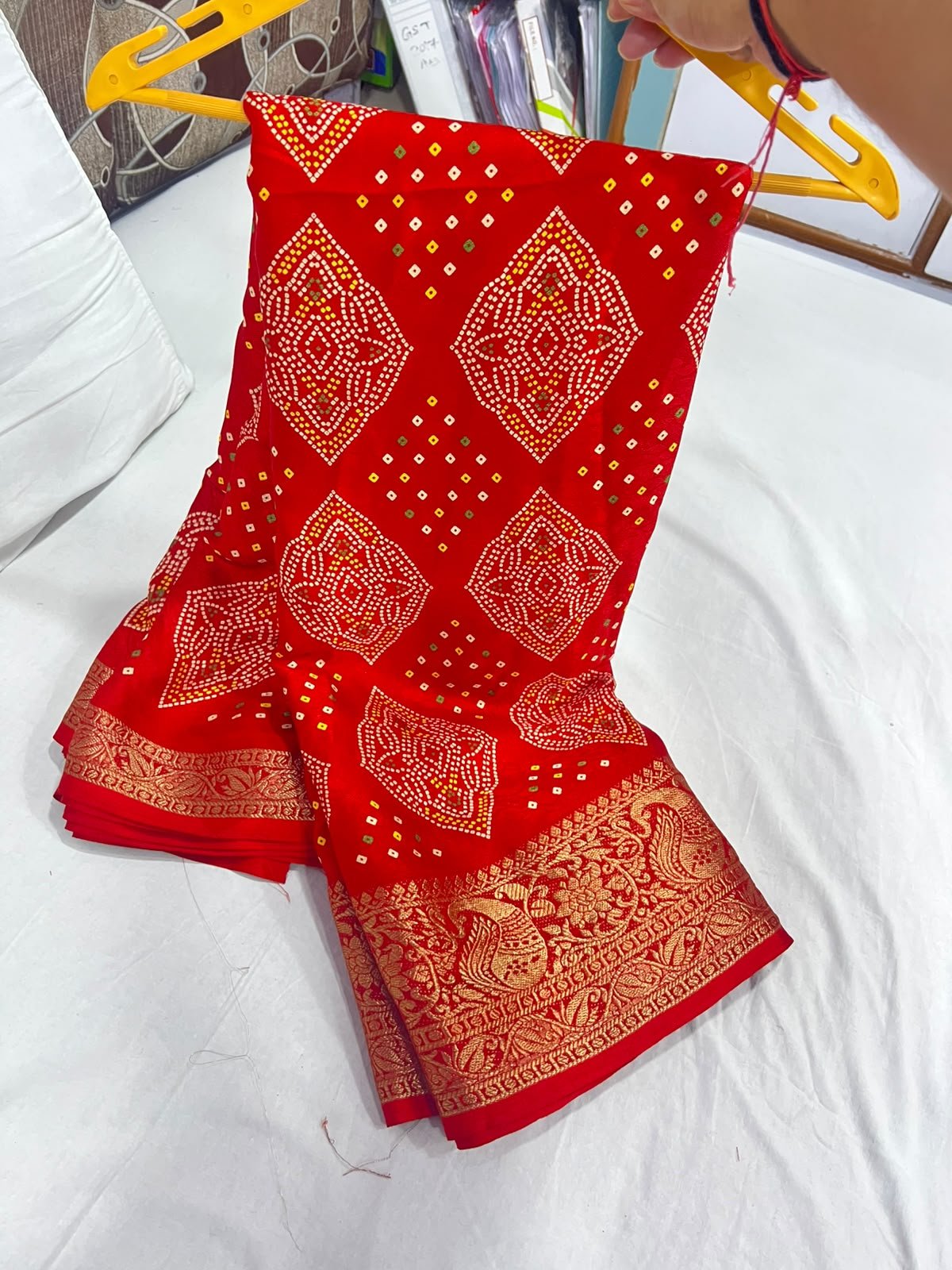 Red Marshmellow Bandhani Saree with Woven Border 1 A vibrant red Marshmellow Bandhani saree with traditional patterns and a wide ornate gold woven border hanging on a yellow hanger.