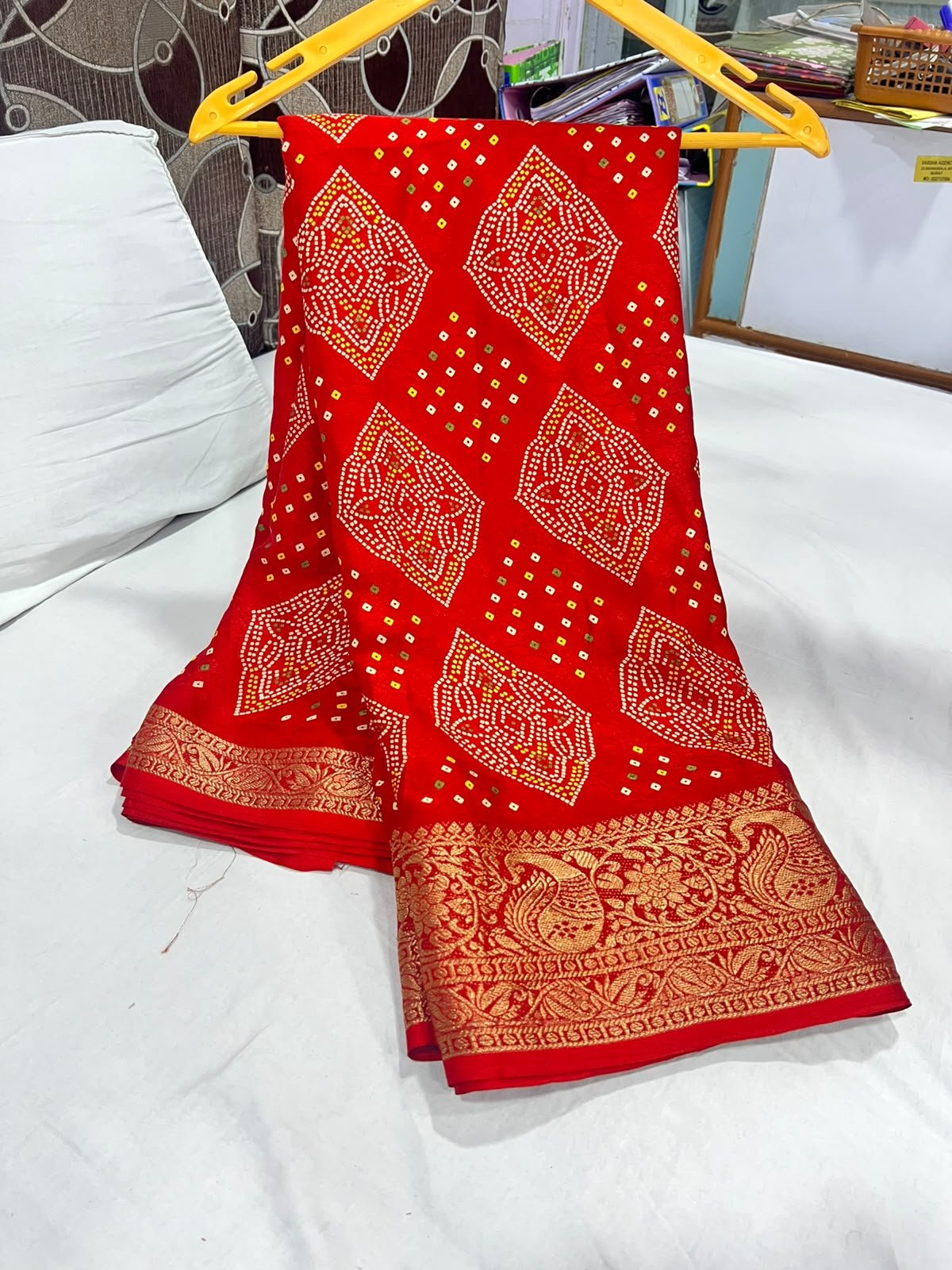 Red Marshmellow Bandhani Saree with Woven Border 2 A vibrant red Marshmellow Bandhani saree with traditional patterns and a wide ornate gold woven border hanging on a yellow hanger.