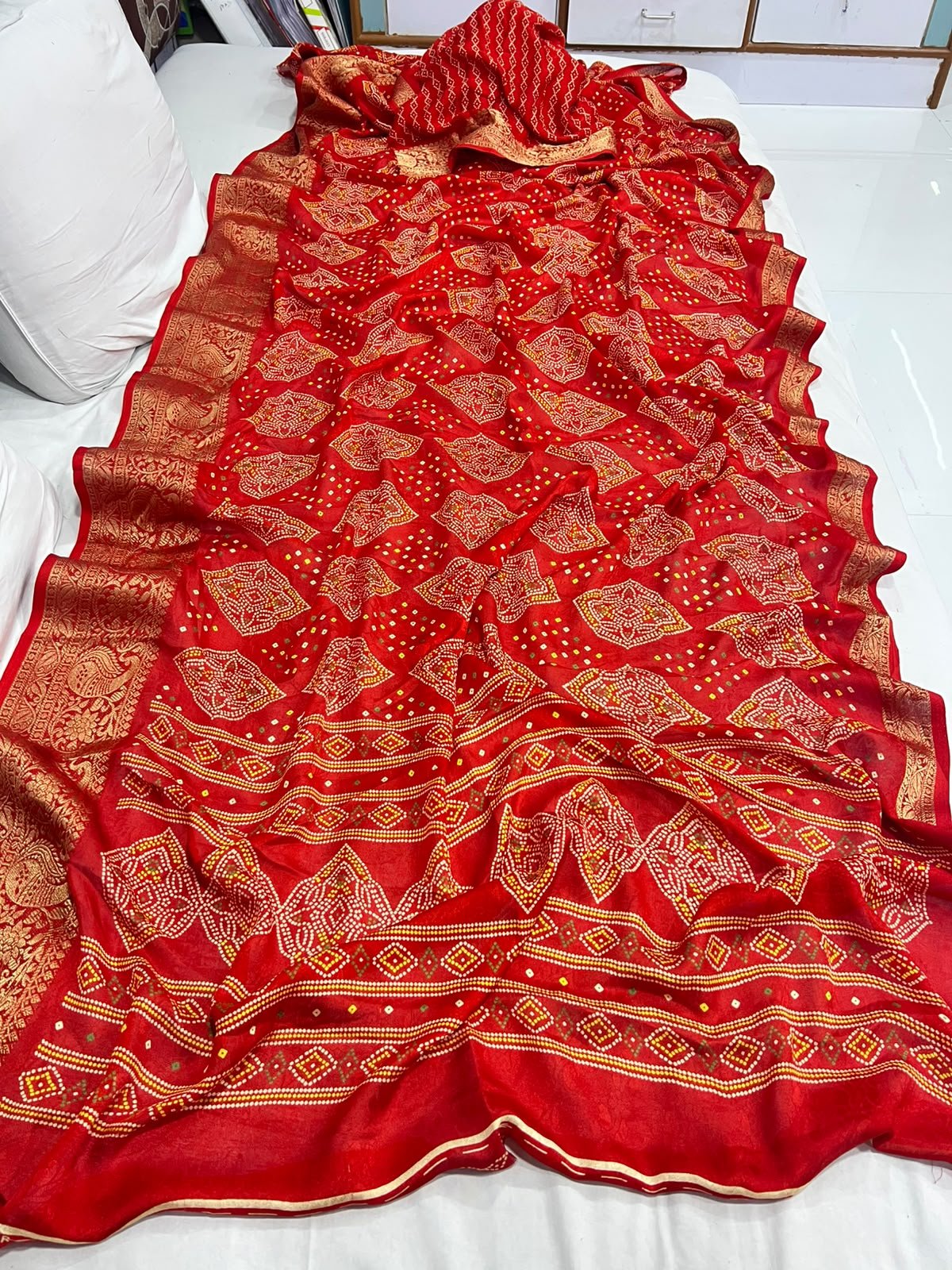 Red Marshmellow Bandhani Saree with Woven Border 3 A vibrant red Marshmellow Bandhani saree with traditional patterns and a wide ornate gold woven border hanging on a yellow hanger.