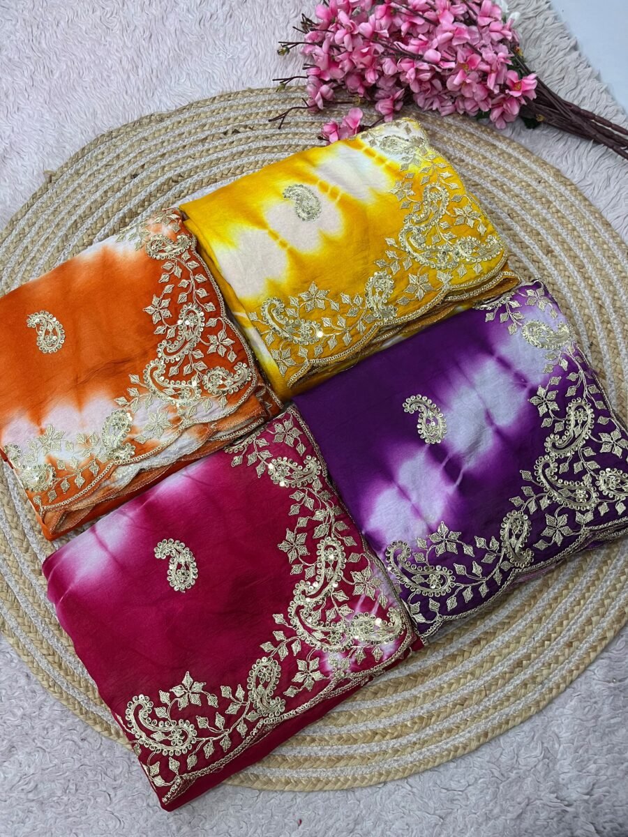 Four vibrant tie-dye sarees in yellow, orange, purple, and maroon, each featuring heavy gold embroidery and cutwork borders, displayed on a circular woven mat.