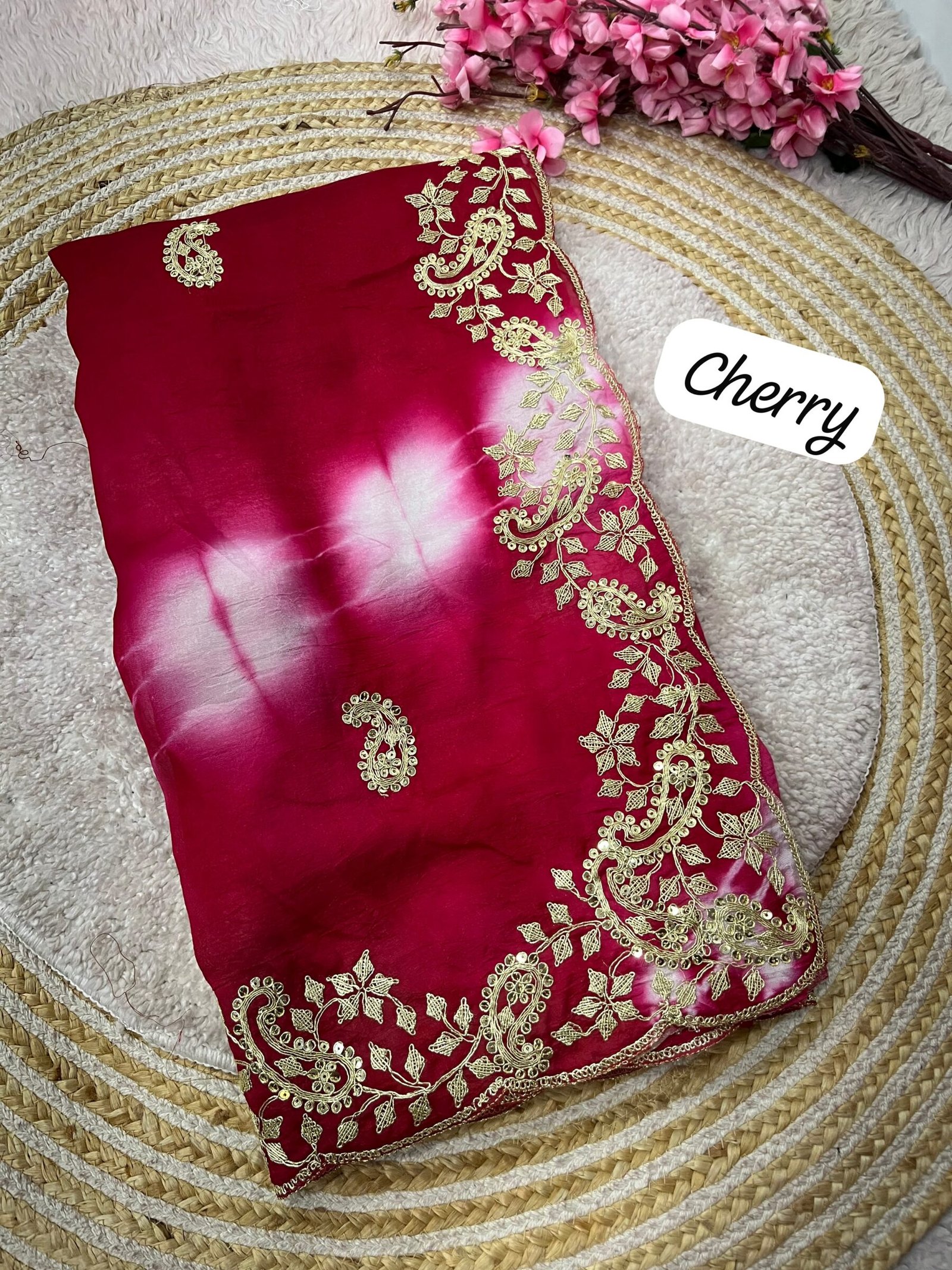 Tana Bana Fabric Saree with Pitta and Sequence Work 2 Four vibrant tie-dye sarees in yellow, orange, purple, and maroon, each featuring heavy gold embroidery and cutwork borders, displayed on a circular woven mat.
