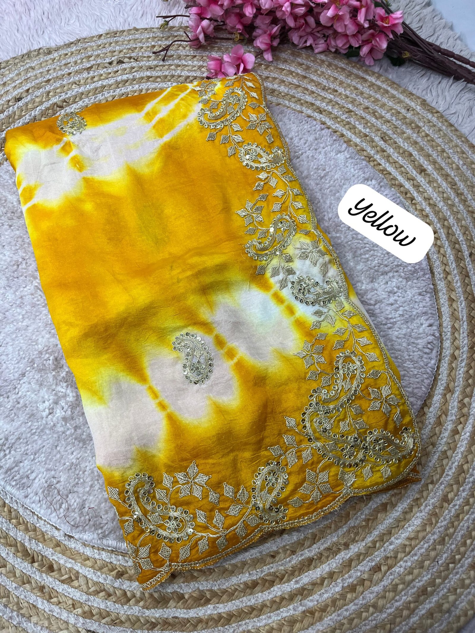 Tana Bana Fabric Saree with Pitta and Sequence Work 3 Four vibrant tie-dye sarees in yellow, orange, purple, and maroon, each featuring heavy gold embroidery and cutwork borders, displayed on a circular woven mat.