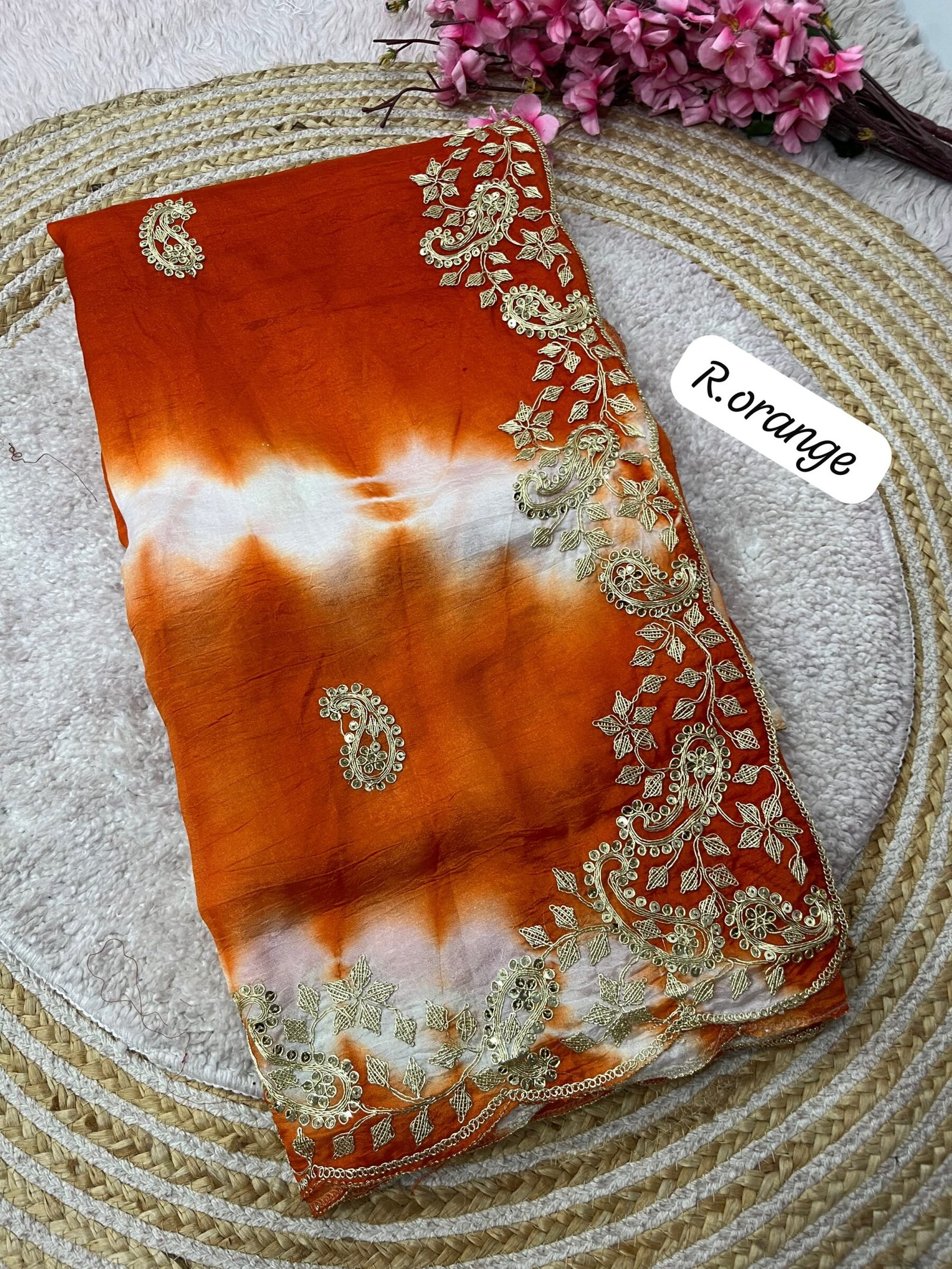 Tana Bana Fabric Saree with Pitta and Sequence Work 4 Four vibrant tie-dye sarees in yellow, orange, purple, and maroon, each featuring heavy gold embroidery and cutwork borders, displayed on a circular woven mat.