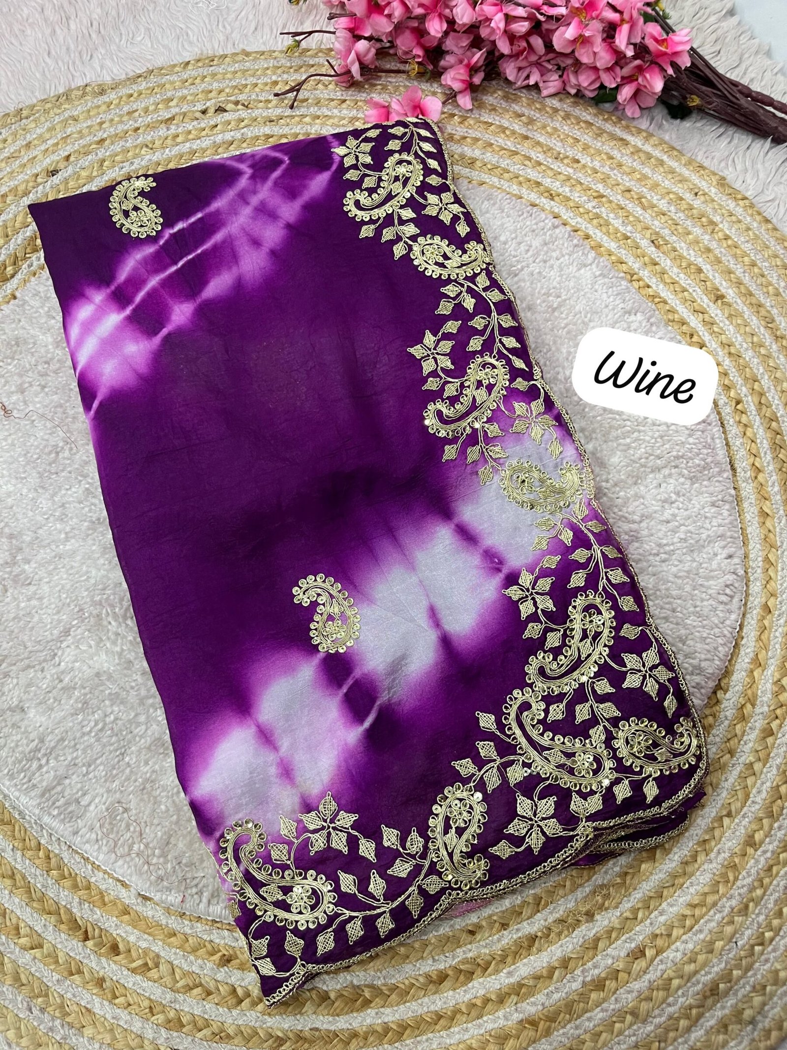 Tana Bana Fabric Saree with Pitta and Sequence Work 5 Four vibrant tie-dye sarees in yellow, orange, purple, and maroon, each featuring heavy gold embroidery and cutwork borders, displayed on a circular woven mat.