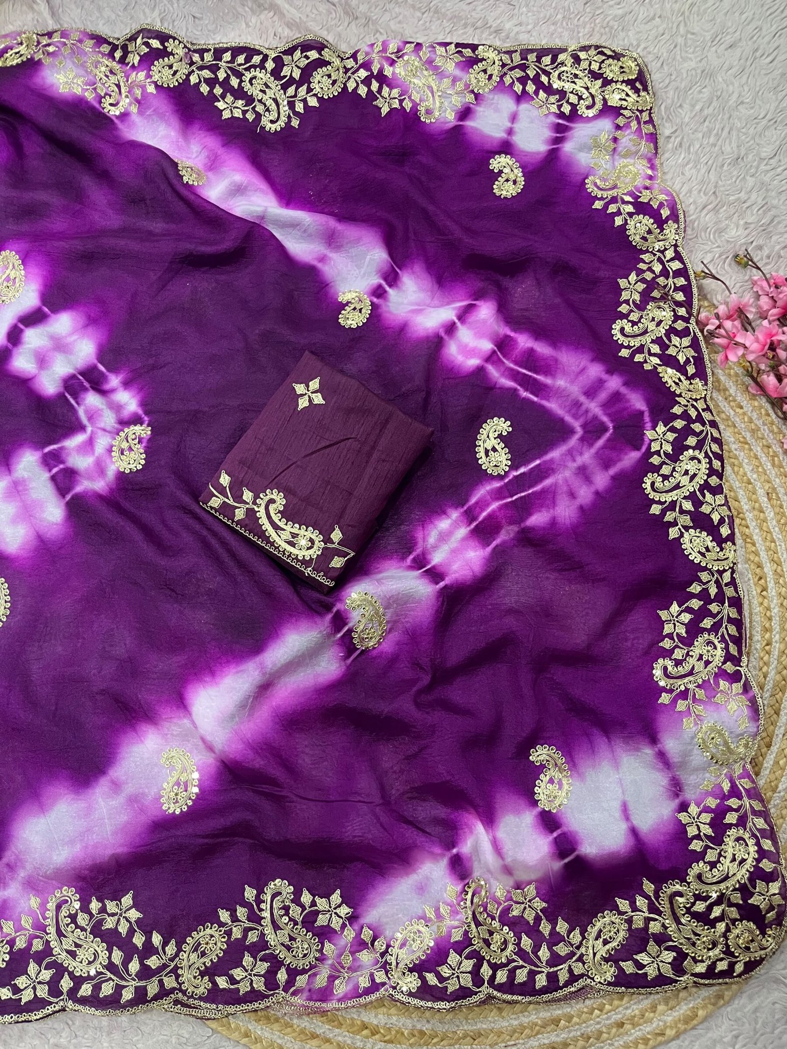 Tana Bana Fabric Saree with Pitta and Sequence Work 7 Four vibrant tie-dye sarees in yellow, orange, purple, and maroon, each featuring heavy gold embroidery and cutwork borders, displayed on a circular woven mat.