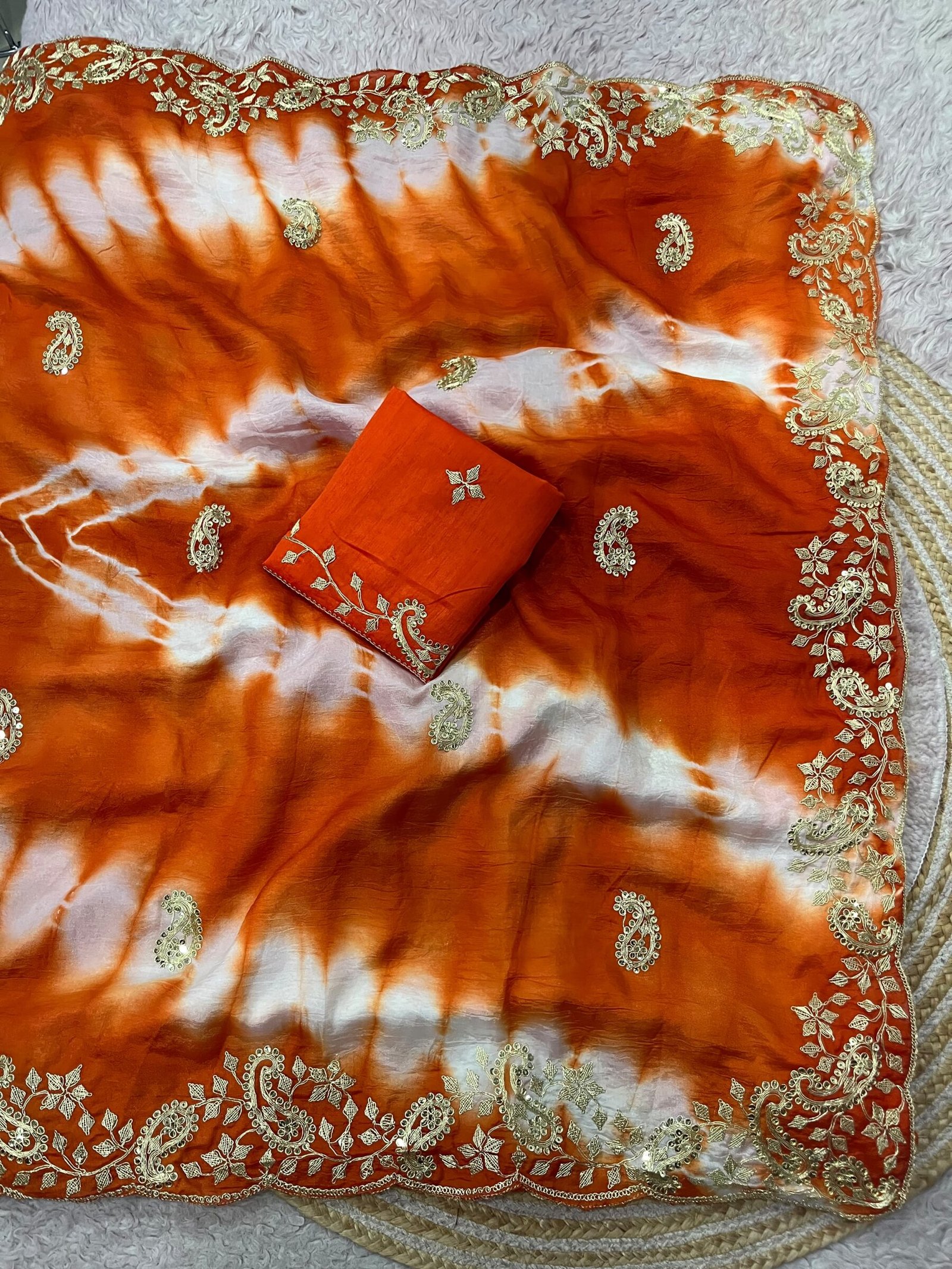 Tana Bana Fabric Saree with Pitta and Sequence Work 8 Four vibrant tie-dye sarees in yellow, orange, purple, and maroon, each featuring heavy gold embroidery and cutwork borders, displayed on a circular woven mat.