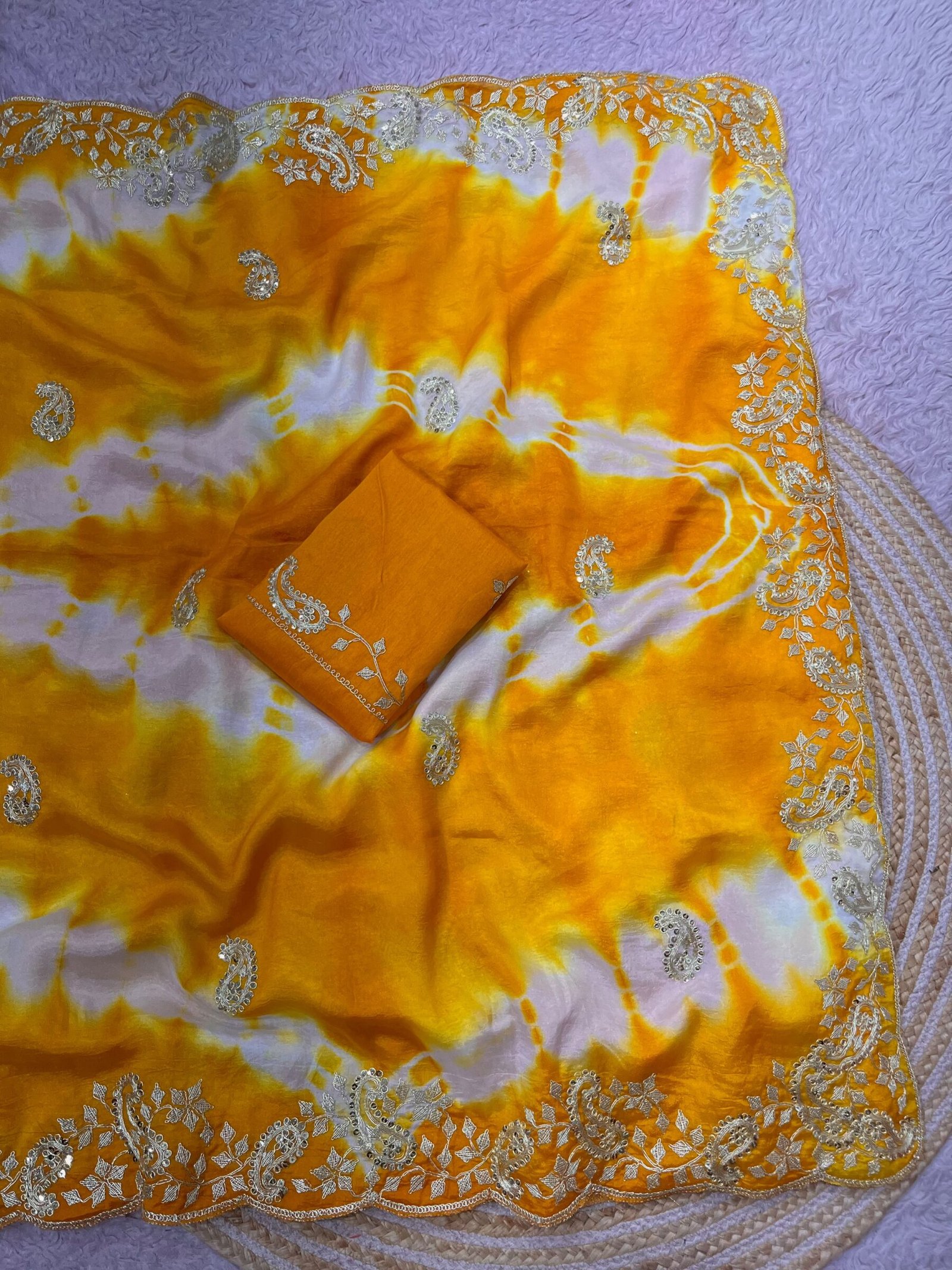 Tana Bana Fabric Saree with Pitta and Sequence Work 9 Four vibrant tie-dye sarees in yellow, orange, purple, and maroon, each featuring heavy gold embroidery and cutwork borders, displayed on a circular woven mat.