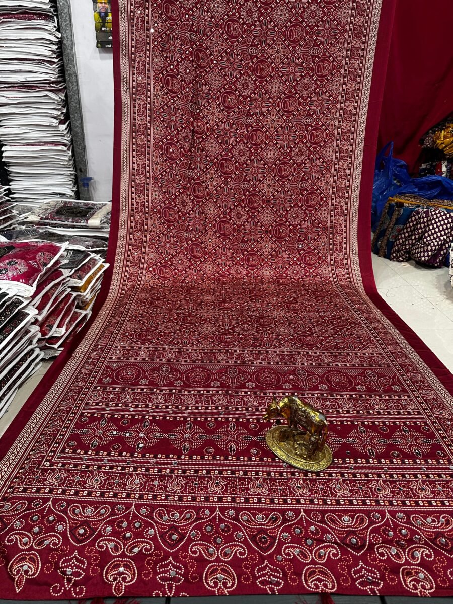 A beautiful deep red Ajarkh Mirror Bandhani saree displayed with a traditional brass cow figurine placed on the fabric.