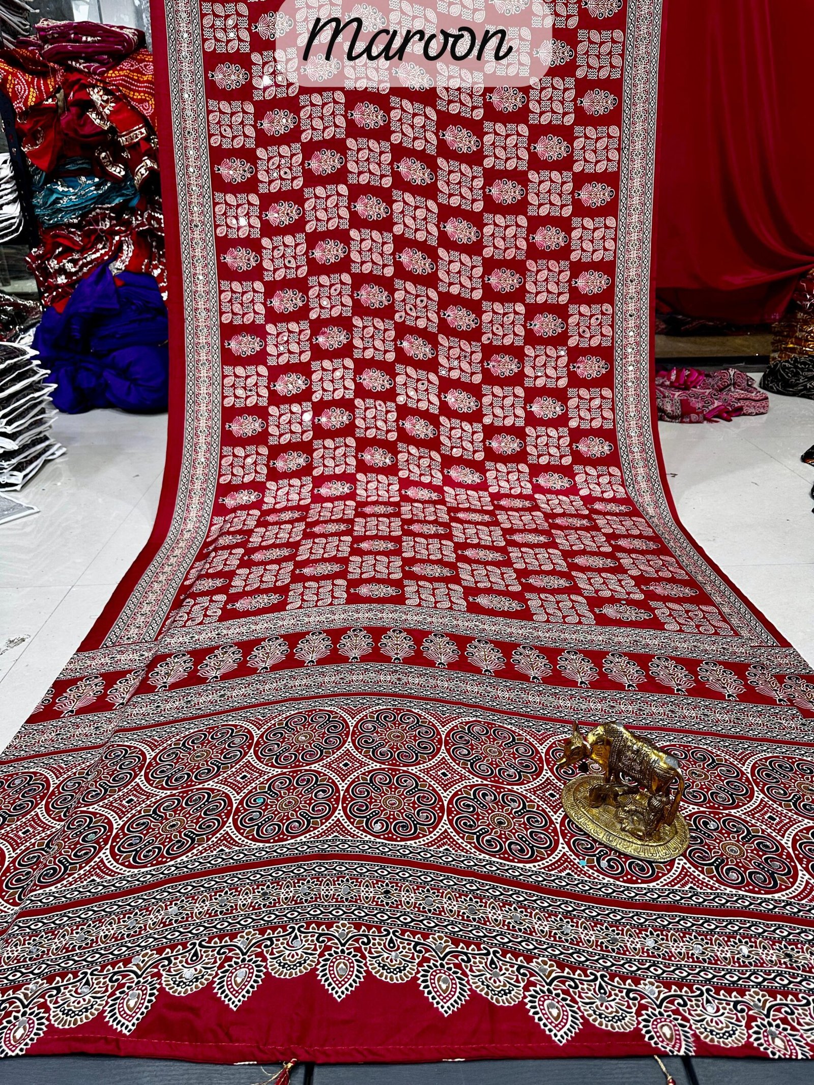 Ajarkh Print Soft Satin Silk Saree with Mirror Work 1 A maroon Ajarkh print soft satin silk saree displayed with a golden statue of a bull, featuring intricate mirror work and traditional patterns.