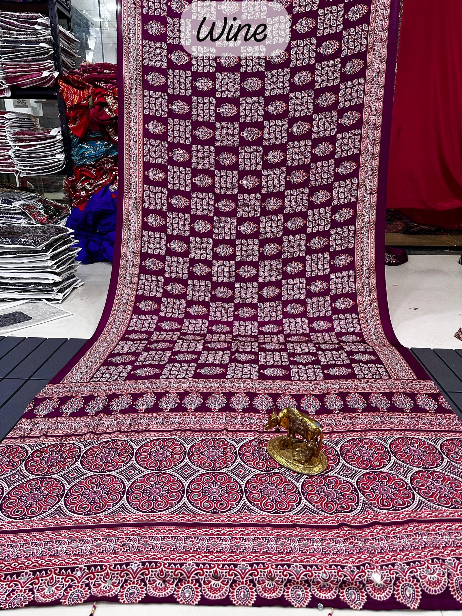 Ajarkh Print Soft Satin Silk Saree with Mirror Work 6 A maroon Ajarkh print soft satin silk saree displayed with a golden statue of a bull, featuring intricate mirror work and traditional patterns.