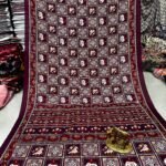 A wine-colored crepe silk saree with Ajrakh print, mirror work, and a matching patterned border displayed on a floor.