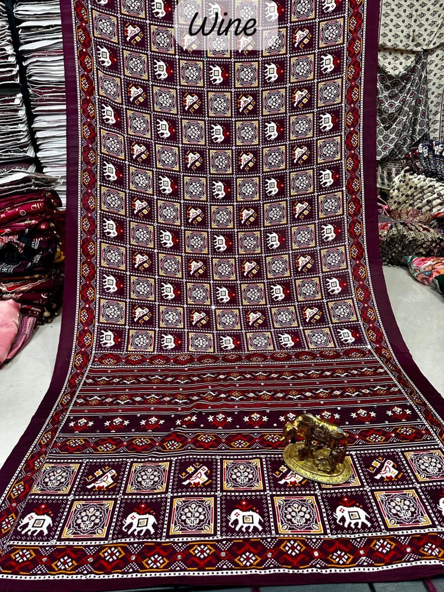 A wine-colored crepe silk saree with Ajrakh print, mirror work, and a matching patterned border displayed on a floor.