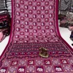 A wine-colored crepe silk saree with Ajrakh print, mirror work, and a matching patterned border displayed on a floor.