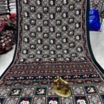 A wine-colored crepe silk saree with Ajrakh print, mirror work, and a matching patterned border displayed on a floor.