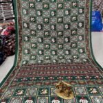 A wine-colored crepe silk saree with Ajrakh print, mirror work, and a matching patterned border displayed on a floor.