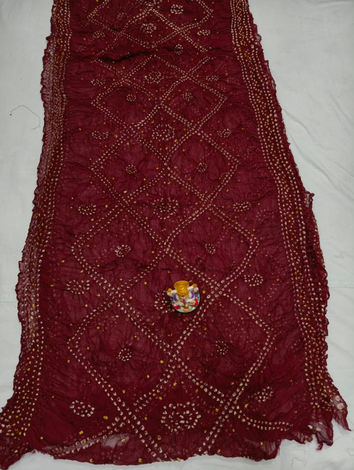 Art Silk Rai Bandhej Saree | Traditional Bandhani Drape 1 A beautiful deep maroon Art Silk Rai Bandhej Saree displayed with a small idol placed on top to highlight its intricate patterned texture.