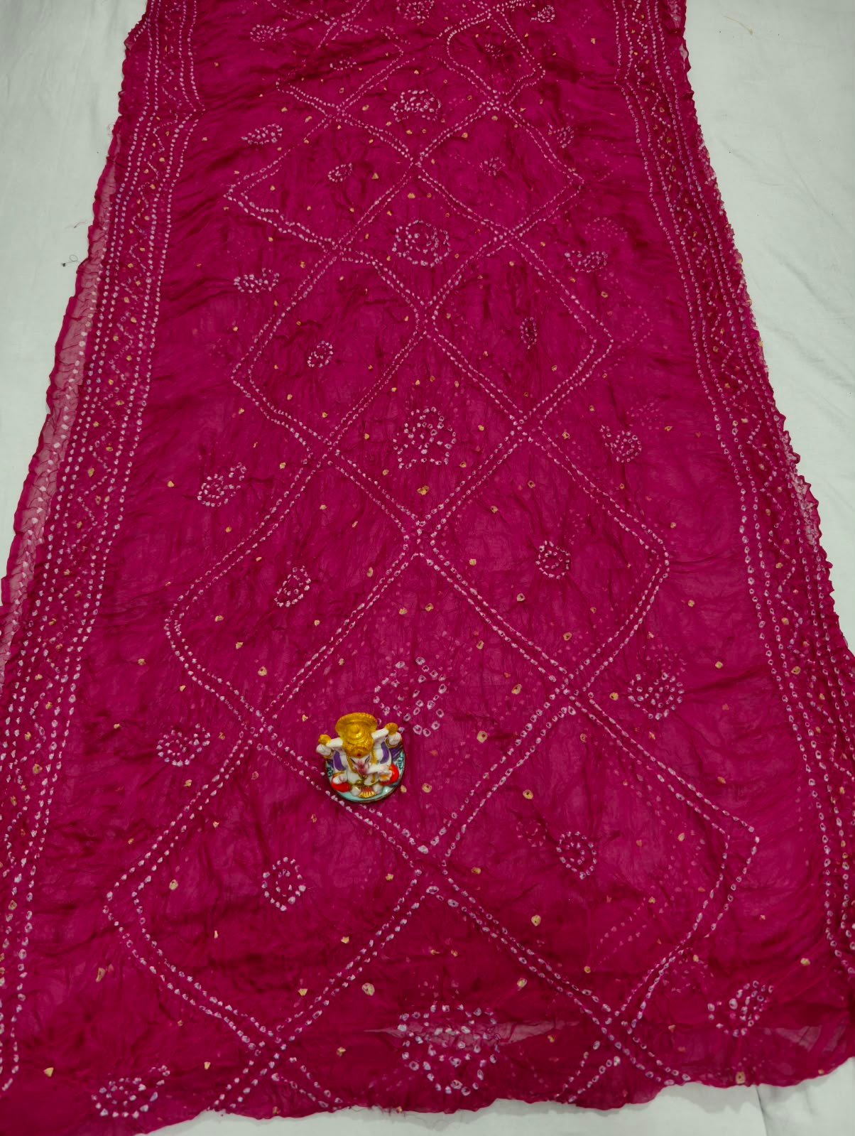 Art Silk Rai Bandhej Saree | Traditional Bandhani Drape 4 A beautiful deep maroon Art Silk Rai Bandhej Saree displayed with a small idol placed on top to highlight its intricate patterned texture.