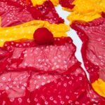 A close-up of a Nazneen Chiffon Bandhani Saree featuring vibrant red and yellow tie and dye patterns.
