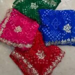 Four vibrant Bandhej sarees in red, pink, blue, and green with heavy Gota Patti borders lying on a floor.