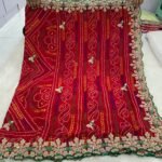 A beautiful red Bandhej saree with heavy coding, sequence work, and a wide embroidered floral border displayed on a white fur background.