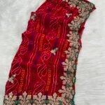 A beautiful red Bandhej saree with heavy coding, sequence work, and a wide embroidered floral border displayed on a white fur background.