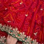 A beautiful red Bandhej saree with heavy coding, sequence work, and a wide embroidered floral border displayed on a white fur background.