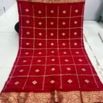 A beautiful red Chinon saree with heavy Zari work, butti, and a golden Jacquard border, being held up to display the design.