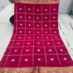 A beautiful red Chinon saree with heavy Zari work, butti, and a golden Jacquard border, being held up to display the design.