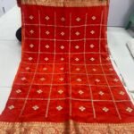 A beautiful red Chinon saree with heavy Zari work, butti, and a golden Jacquard border, being held up to display the design.