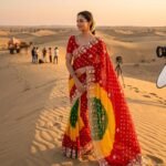 A woman posing in the desert wearing a vibrant red, yellow, and green Chunri Bandhani saree with intricate pita work.