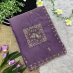 A beautiful teal designer saree laid out flat with peacock embroidery and sequin work, surrounded by white flowers and purple tulips.