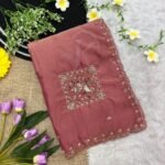 A beautiful teal designer saree laid out flat with peacock embroidery and sequin work, surrounded by white flowers and purple tulips.