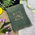 A beautiful teal designer saree laid out flat with peacock embroidery and sequin work, surrounded by white flowers and purple tulips.