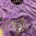 A beautiful teal designer saree laid out flat with peacock embroidery and sequin work, surrounded by white flowers and purple tulips.