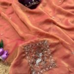 A beautiful teal designer saree laid out flat with peacock embroidery and sequin work, surrounded by white flowers and purple tulips.