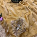 A beautiful teal designer saree laid out flat with peacock embroidery and sequin work, surrounded by white flowers and purple tulips.