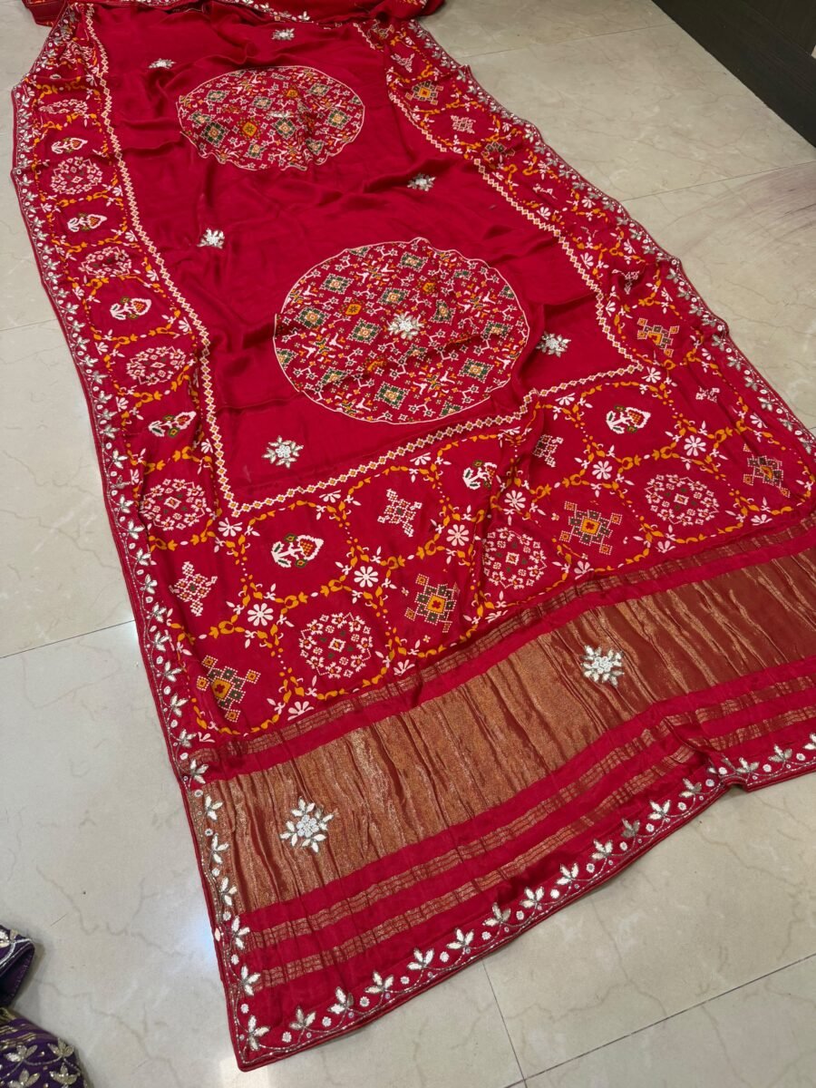 A beautiful red pure Modal Gajji Patola saree with intricate Gota Patti handwork and a metallic zari border spread across a tiled floor.